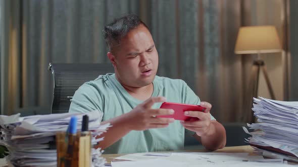 Close Up Of Fat Asian Man Being Upset Losing Game On Smartphone After Working With Documents