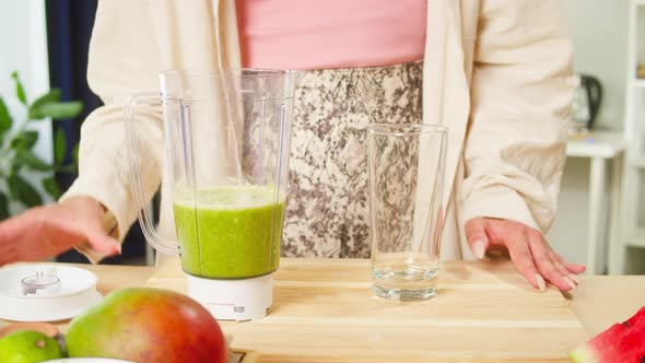 Young Woman Opening Blender and Pouring Green Detox Smoothie Into Glass on Table in the Kitchen