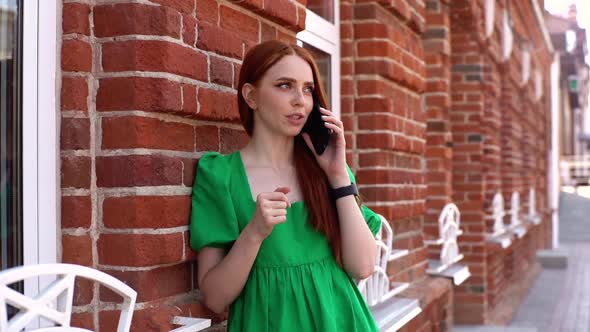 Young Smiling Woman Talking on Mobile Phone Standing Leaning on Old Stone Wall of Building in Town