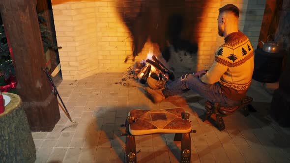 Young bearded guy sit on the chair, rubbing his hands to warm up near fire. 