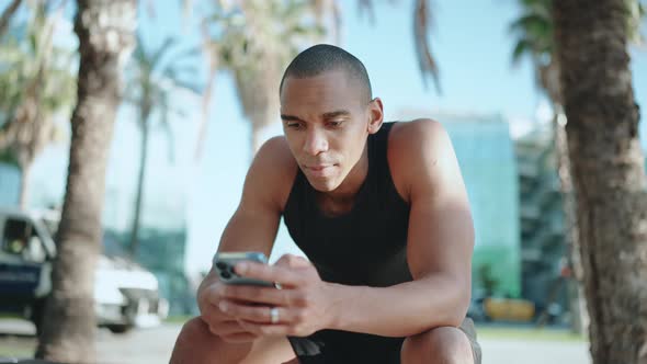 Bald pretty sportsman wearing black T-shirt typing by phone on the bench