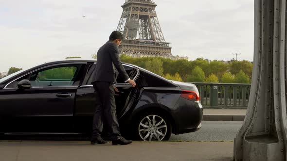 Driver in Suit Opens the Car Door for a Client on the Background of Eiffel Tower