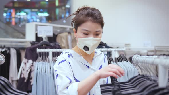 asian female shopper wearing facial virus protective mask choosing clothes at the store