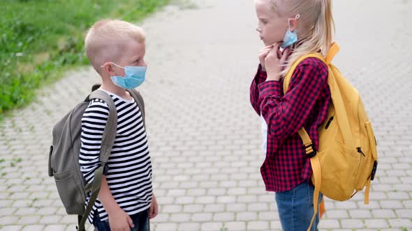 Caucasian Children in Mask Blonde Sister Straightens Brother Face Mask Kids with Backpacks on Street