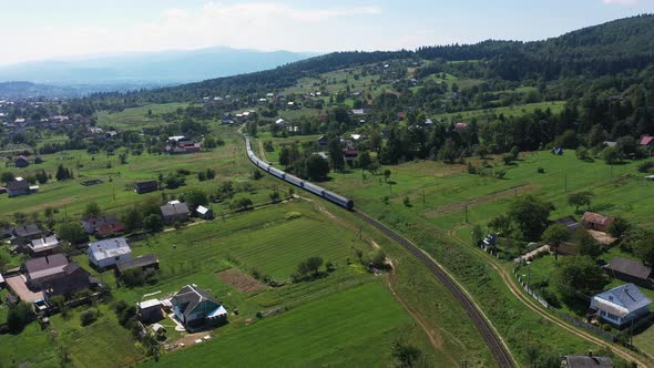 Train Moving on Railroad Tracks in Carpathian Mountains Through the Village Aerial View