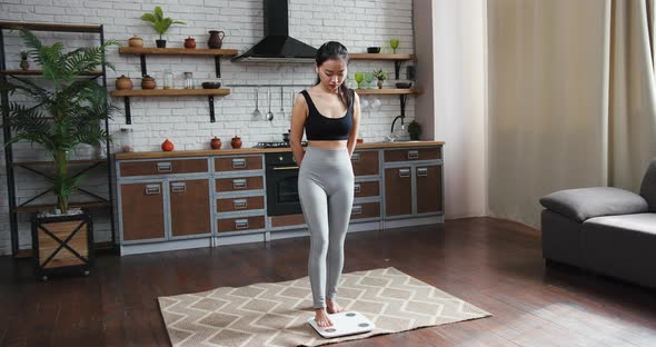 Asian Woman in Sportswear Standing on Weight Scale and Rejoicing By Weight Loss in Home Interior