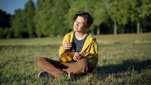 Talented Confident Romantic Teenage Boy Sitting on Sunny Summer Lawn Playing Ukulele