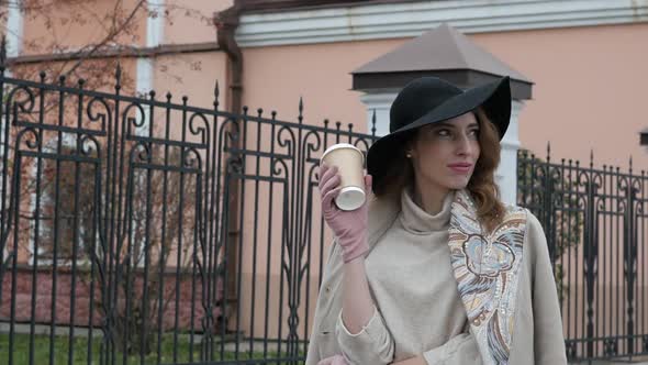 Young Redhaired Woman in a Coat and a Black Hat on the Background of the City Drinks Coffee