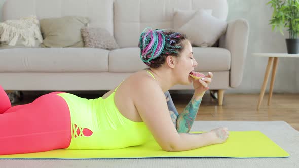 Plump Young Woman in Sportswear Enjoying of Eating Donut Lying on Mat at Home