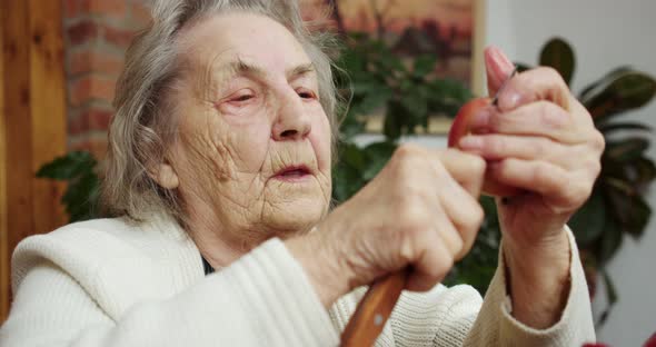 an Elderly Grandmother Cleans a Red Apple with a Knife Sitting at Home and Enthusiastic About the