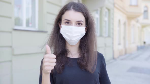 A Young Turkish Woman in a Face Mask Shows a Thumb Up To the Camera in the Street in an Urban Area