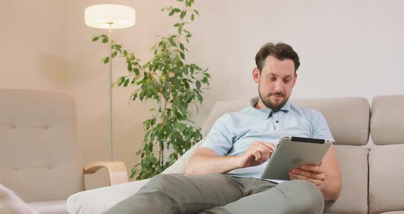 Young Bearded Man is Lying on the Sofa and Holding Tablet in Hands He is Relaxing and Scrolling the