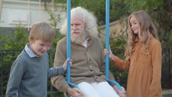 Middle Shot of Happy Caucasian Brother and Sister Swinging Grandfather on Swings. Portrait of
