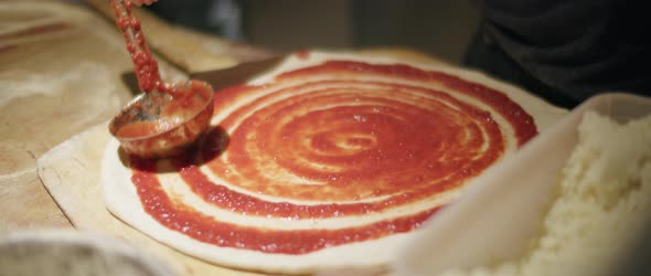 Chef spreading tomato sauce with metal ladle on pizza dough base