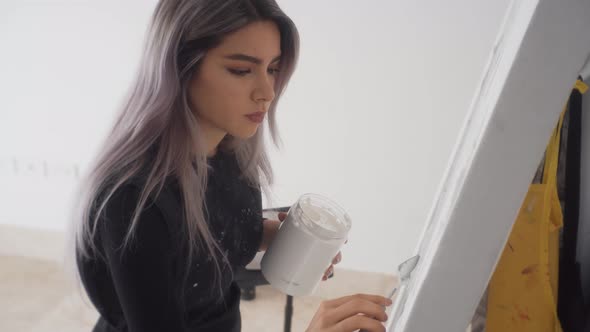 Young Beautiful Painter Standing And Applying White Acrylic Paint On Canvas In Easel. - medium shot