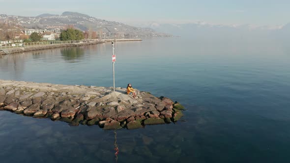 Aerial of woman sitting at the end of quay at beautiful lake with mountains in the far background