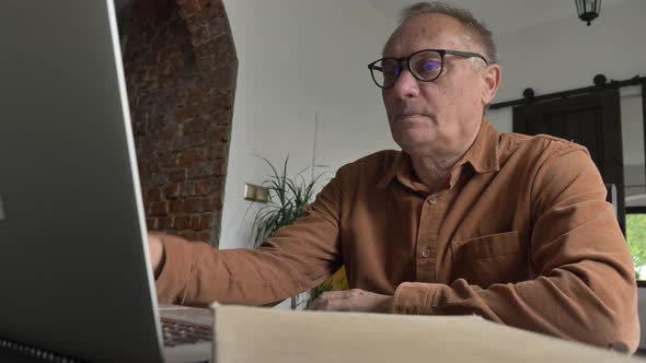 Old man in eyeglasses using laptop computer at home.