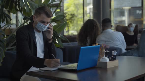 Portrait of Concentrated Caucasian Businessman in Covid-19 Face Mask Talking on the Phone in Cafe