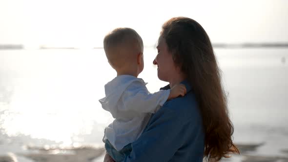 A Young Caucasian Woman Hugs a Toddler Near the Ocean at Dawn