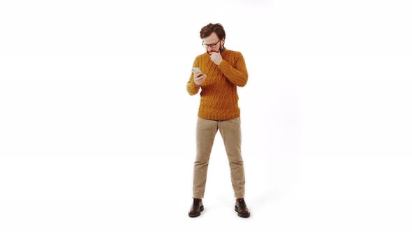 Young Adult Caucasian Guy in Glasses Looking at His Cellphone Contemplating and Touching His Beard