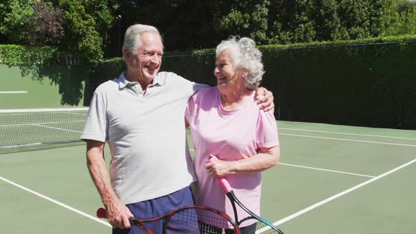 Happy senior caucasian couple holding tennis rackets and embracing in tennis court