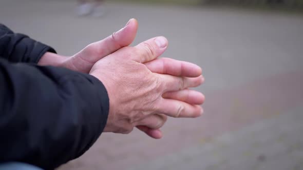 Seated Male Rubs Warm Hands Outdoors in the Park