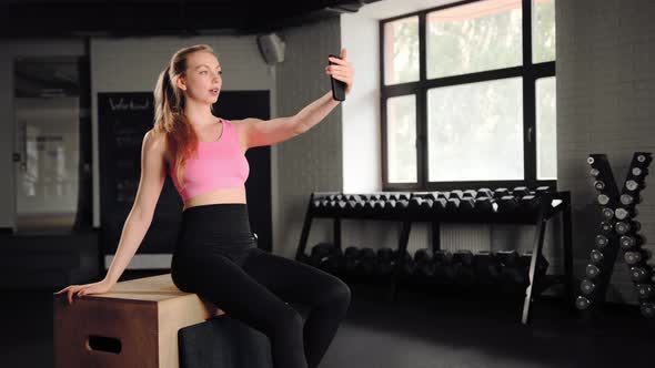 Athlete girl is resting after exercise with the phone while sitting in the gym.