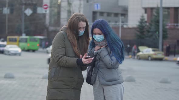 Brunette Woman in Protective Mask Showing Latest News About Coronavirus To Friend on Smartphone