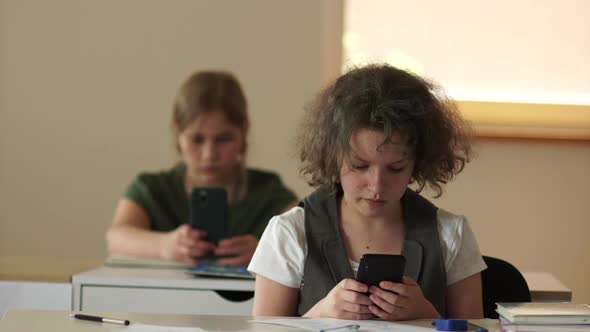 Two Schoolgirls are Sitting at Their Desks at School and Looking at Their Smartphones