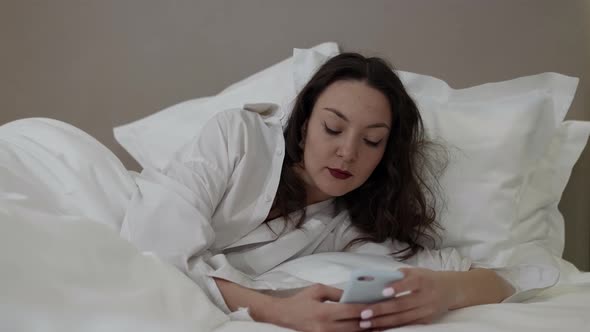 A Pensive Brunette with Bright Lipstick in a White Shirt Lies and Rests in the Room on White Bed