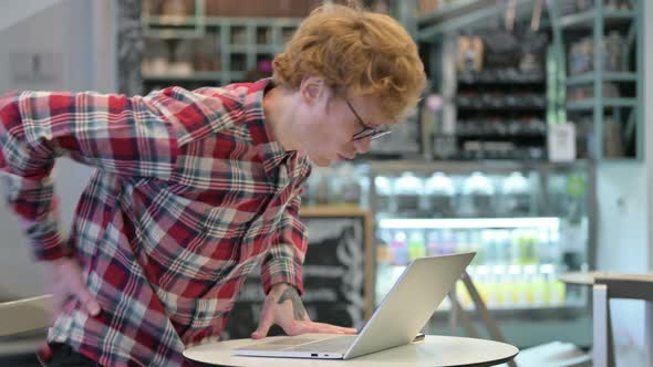 Young Redhead Man with Back Pain Using Laptop in Cafe 