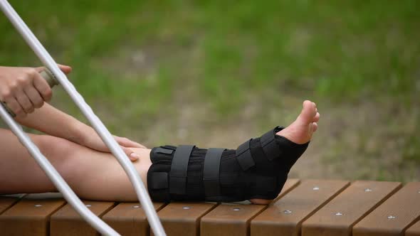 Female With Crutch Sitting Bench With Ankle Brace on Leg, Bone Fracture, Strain