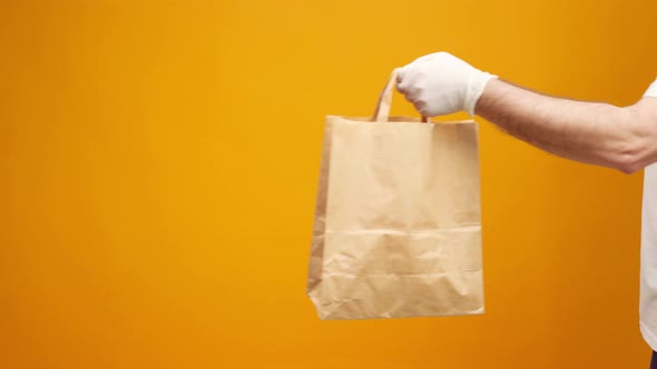 Courier in Gloves Passing Craft Shopping Bag with Delivery Against Yellow Background