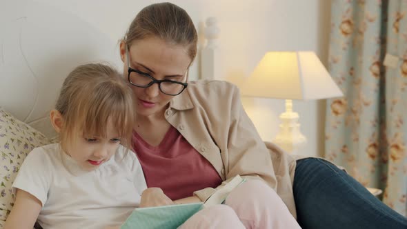 Young Woman Reading Book To Little Girl Relaxing in Bed in Modern Apartment