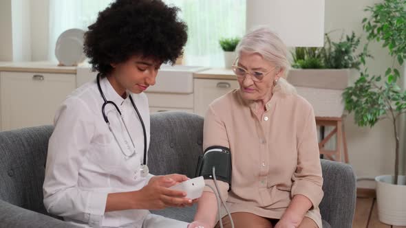 African American Woman Doctor Checking Blood Pressure of Senior Woman While Sitting on Couch at Home