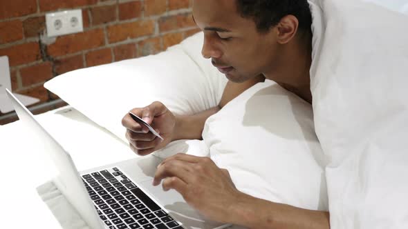 Online Shopping by African Man in Bed, Paying by Bank Credit Card