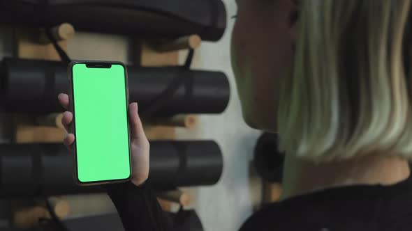 A Young Athletic Woman Holds A Smartphone in Her Hand, Shows A Green Chromakey Screen, Is in The Gym