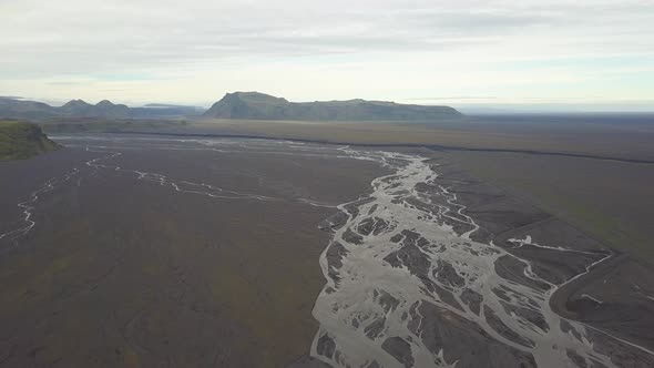 Aerial trucking of stream of water in lowland fiord area, verdant hills in background on an overcast