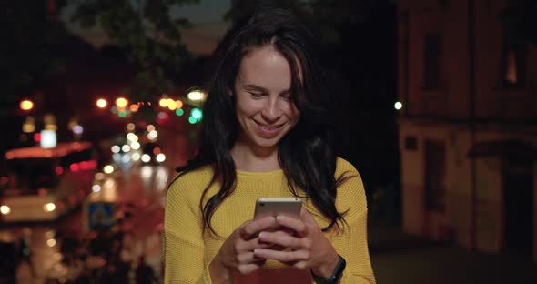 Brunette in Yellow Sweater which Using Her Smartphone in the Evening Among Lights of City Streets