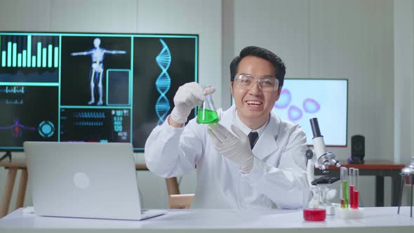 Young Asian Male Research Scientist Holding Glass Test-Tube And Speaking To The Camera