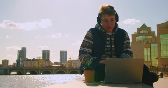 Teen Blogger During an Online Video Broadcast  He is Sitting on Embankment with Laptop