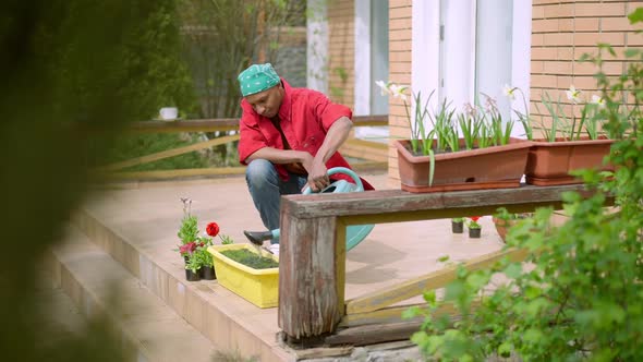 Focused African American Man Watering Soil with Water Sitting in Squat on Wooden Porch