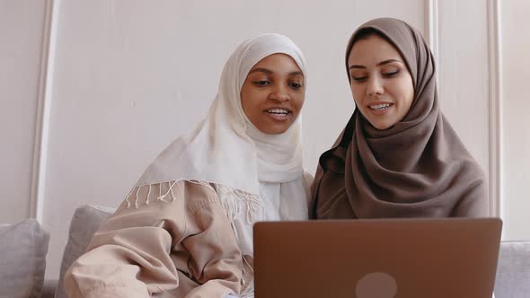 Two Muslim Girls Look at the Laptop Smiling and Laughing in Bright Beige Studio