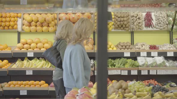 Side View of Two Blond Caucasian Housewives Walking Along Rows in Grocery and Chatting. Serious