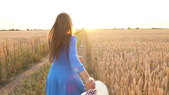 Beautiful Woman in a Blue Dress and Hat Runs Through a Wheat Field at Sunset