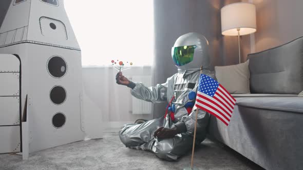 African Man in an American Astronaut Costume Sitting on the Floor in Living Room at Home and Playing