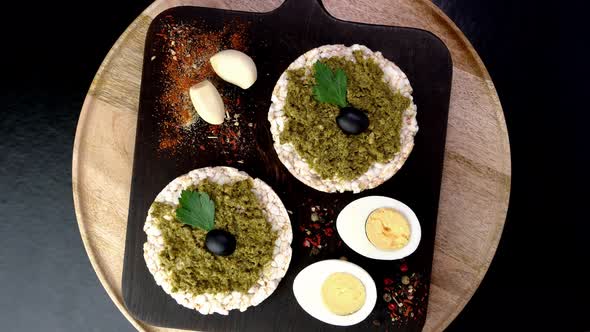 Rice cake toasts with spreading green pesto sauce and boiled eggs rotate slowly on cutting board.