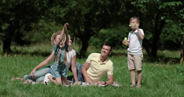 Family Day, Picnic in Nature, the Boy Plays and Blows Soap Bubbles, Young Parents and Daughter Sit