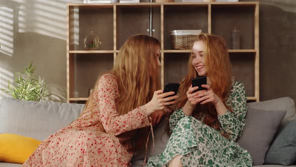 Two Young Twins Sisters Use Smartphones on Sofa in the Living Room