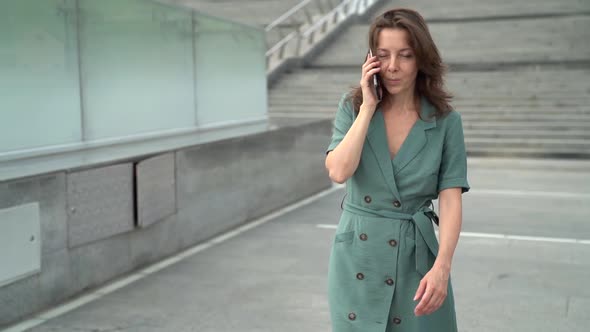 A Middle-aged Woman in a Button-down Dress Walks and Talks on a Smartphone on a City Street on a